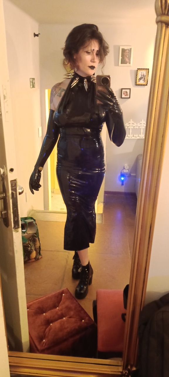 A selfie of a woman, in front of a mirror, wearing latex dress and gloves.